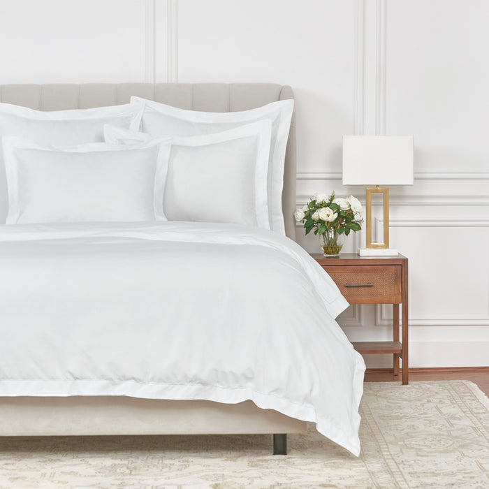 A white cotton duvet cover displayed on a bed with a matching white pillowcases, each finished with hemstitching, in a room with a wooden nightstand, a lamp, and a floral arrangement.